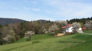 A white house with a red roof on a grassy hill, surrounded by trees and a forested hill in the background. - Ankommen, Entspannen & Genießen - Gemütliches Apartment mit Traumhaftem Ausblick (Lalling)