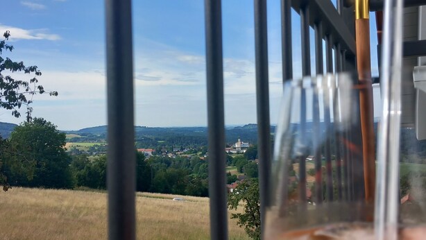 A balcony with a glass door, a glass of amber liquid, and a view of a rural landscape with fields and a distant village. - Ankommen, Entspannen & Genießen - Gemütliches Apartment mit Traumhaftem Ausblick (Lalling)