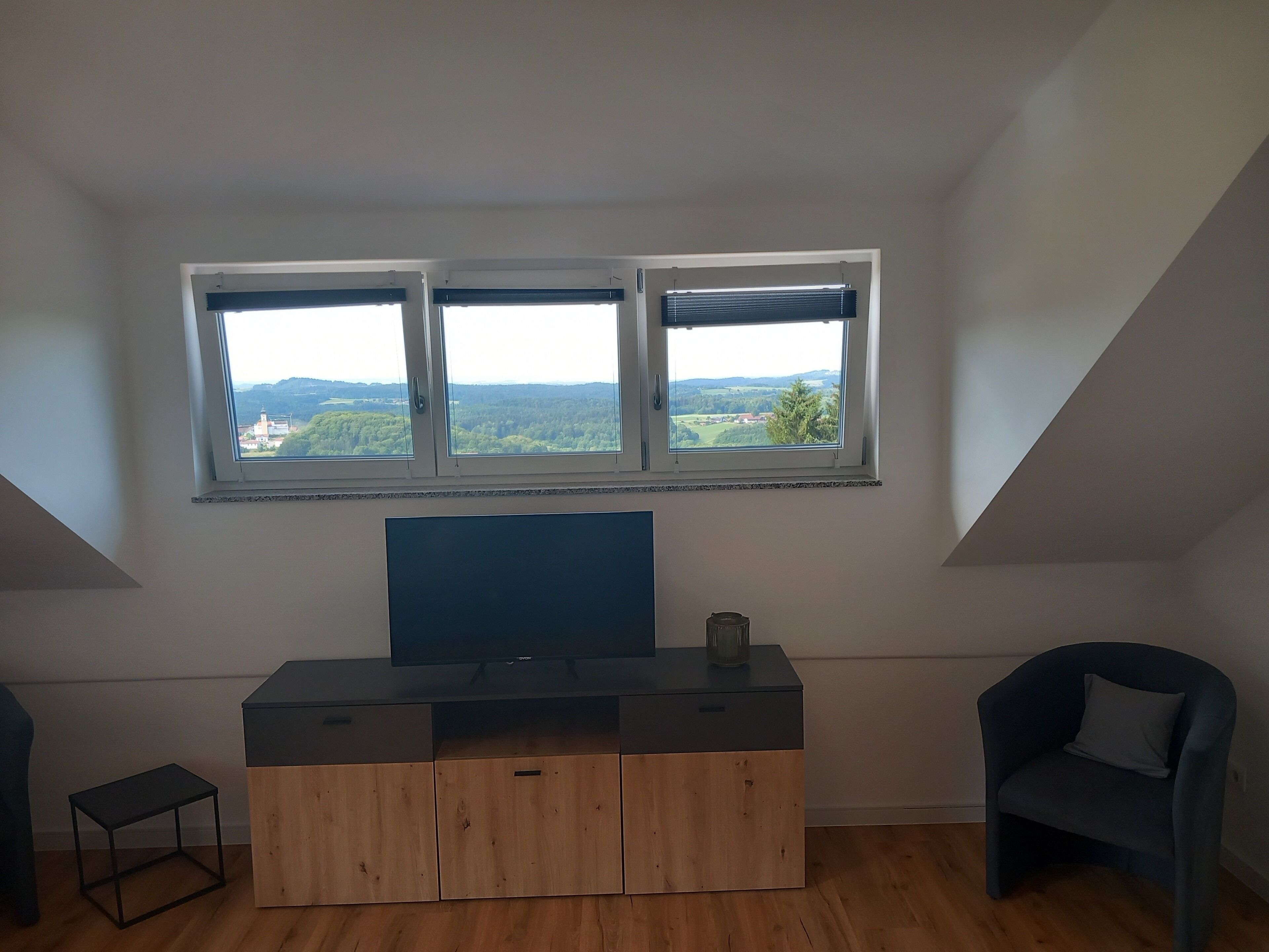 Een kamer met een grote televisie op een houten kast, drie ramen met uitzicht op het platteland, en een donkere fauteuil.