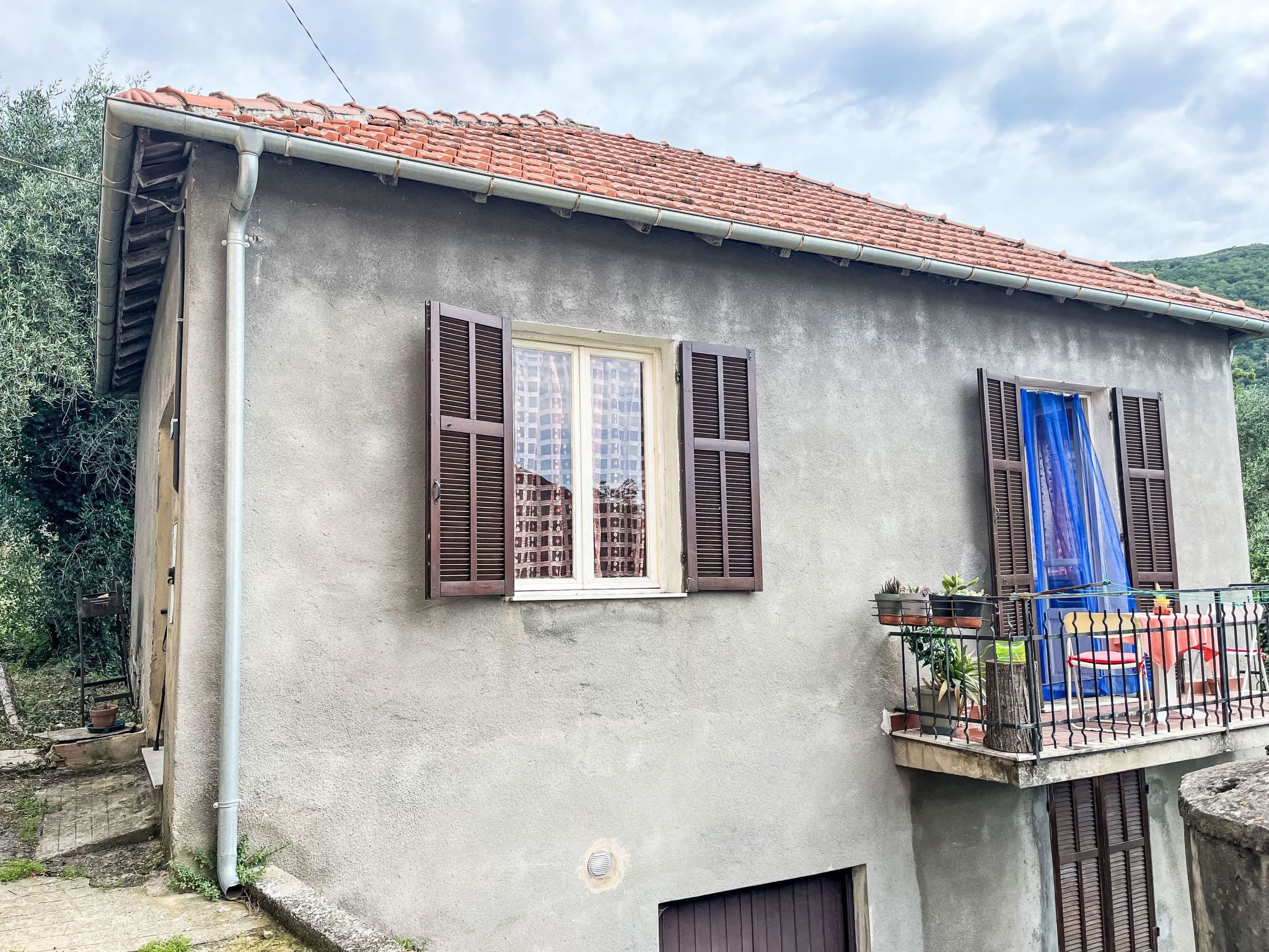 A two-story building with a red-tiled roof, a balcony with potted plants, and a window with brown shutters.