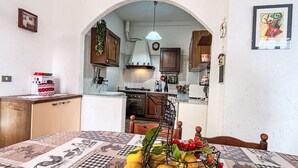 A kitchen with an archway leading to a dining area. There is a table with a patterned tablecloth, a fruit basket, and a stove.