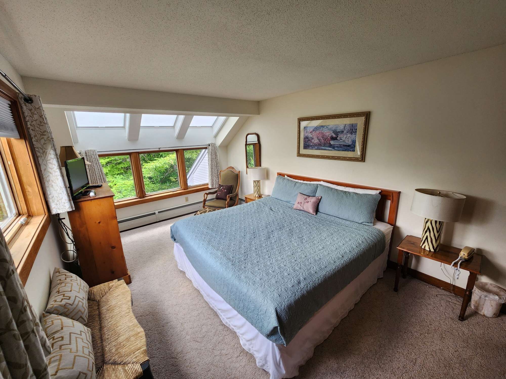 A bedroom with a large bed, a desk with a computer, a chair, a window with curtains, and a painting on the wall.