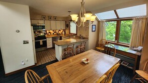 A compact kitchen with a dining area, featuring a wooden table and chairs, a chandelier, and a window with a view of greenery. - Ski In/Out Sunrise West Glade 3 BR 3 BA Trailside, Sauna, Pool, Hot Tub (Killington)