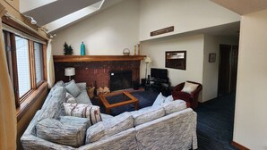A living room with a fireplace, a sofa, a red armchair, and a television. - Ski In/Out Sunrise West Glade 3 BR 3 BA Trailside, Sauna, Pool, Hot Tub (Killington)