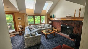 A cozy living room with a sectional sofa, a fireplace, and a view of the outdoors through large windows. - Ski In/Out Sunrise West Glade 3 BR 3 BA Trailside, Sauna, Pool, Hot Tub (Killington)