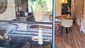 Ein modernes Zimmer mit Glastrennwand, einem Bett mit grauer Bettwäsche, einem kleinen runden Tisch mit einer Vase und einer Pendelleuchte.