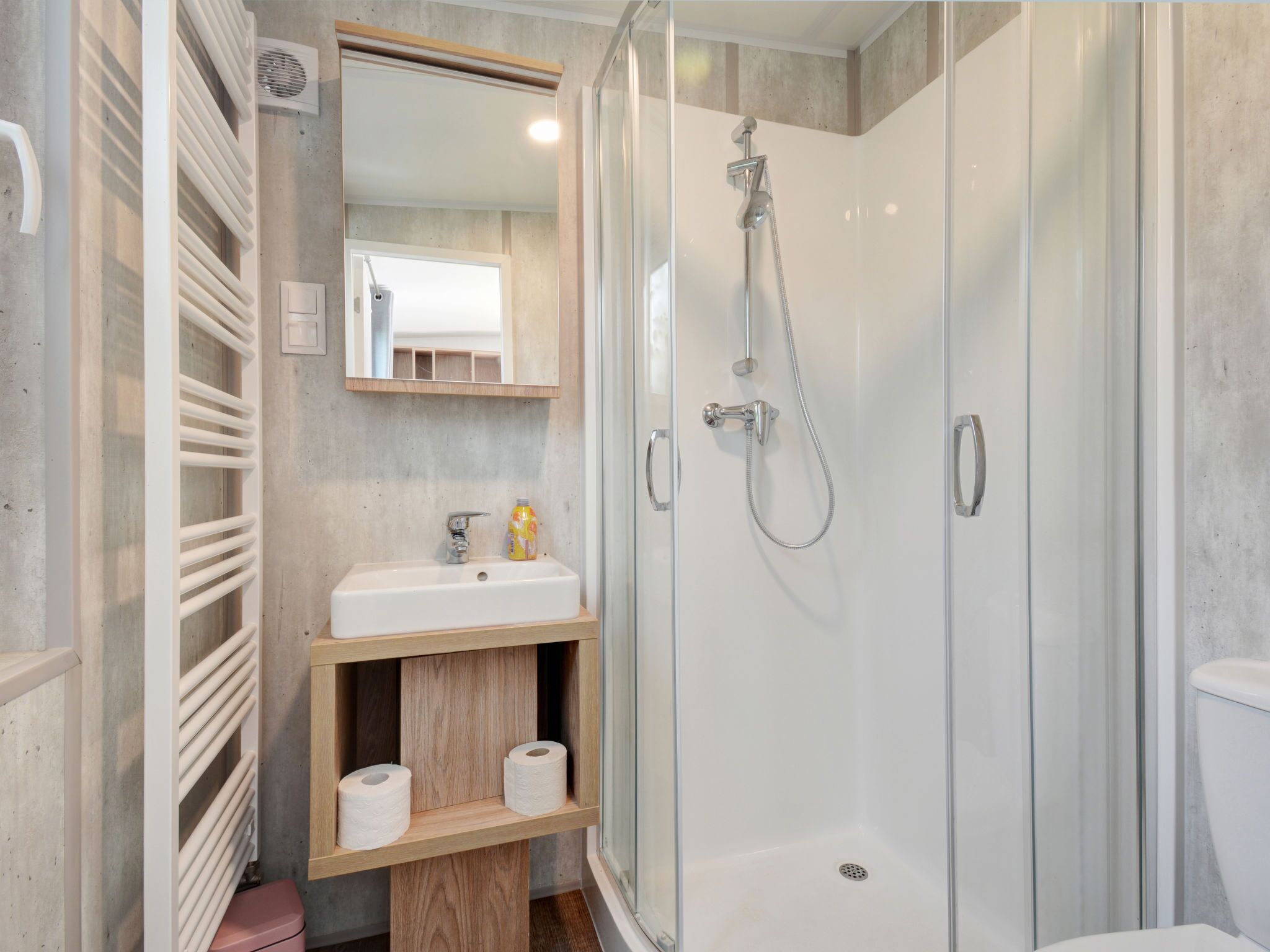 A modern bathroom with a glass-enclosed shower, a white sink, and a mirror above it. There are wooden shelves below the mirror holding toilet paper and a bottle.