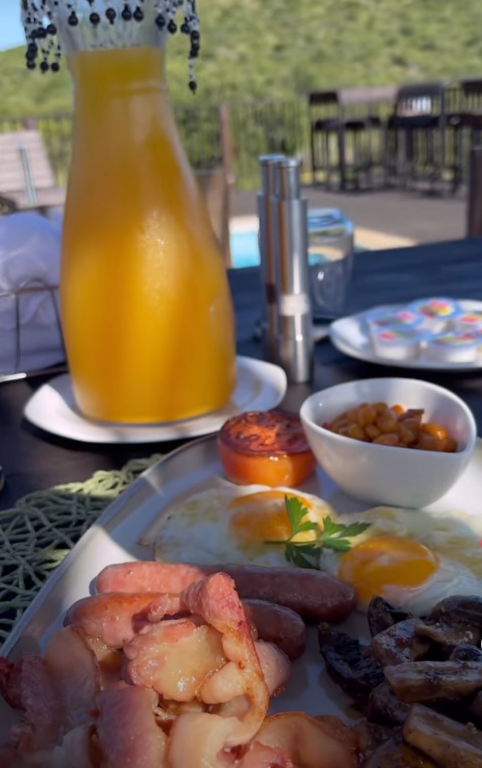 A breakfast setting with a plate of eggs, sausages, and bacon, a glass of orange juice, and a bowl of fruit.