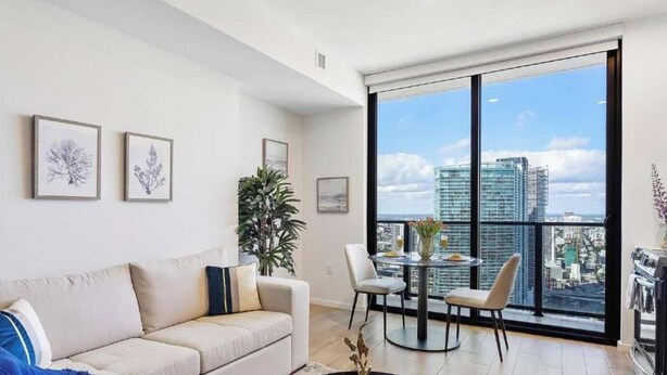 A modern living room with a large window, a sofa, a dining table with chairs, and framed artwork on the wall.