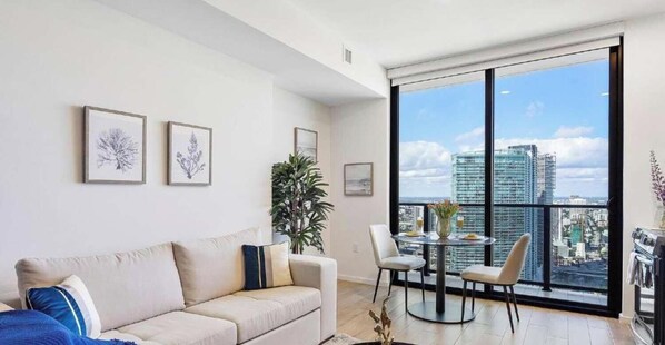 A modern living room with a large window, a sofa, a dining table with chairs, and framed artwork on the wall.