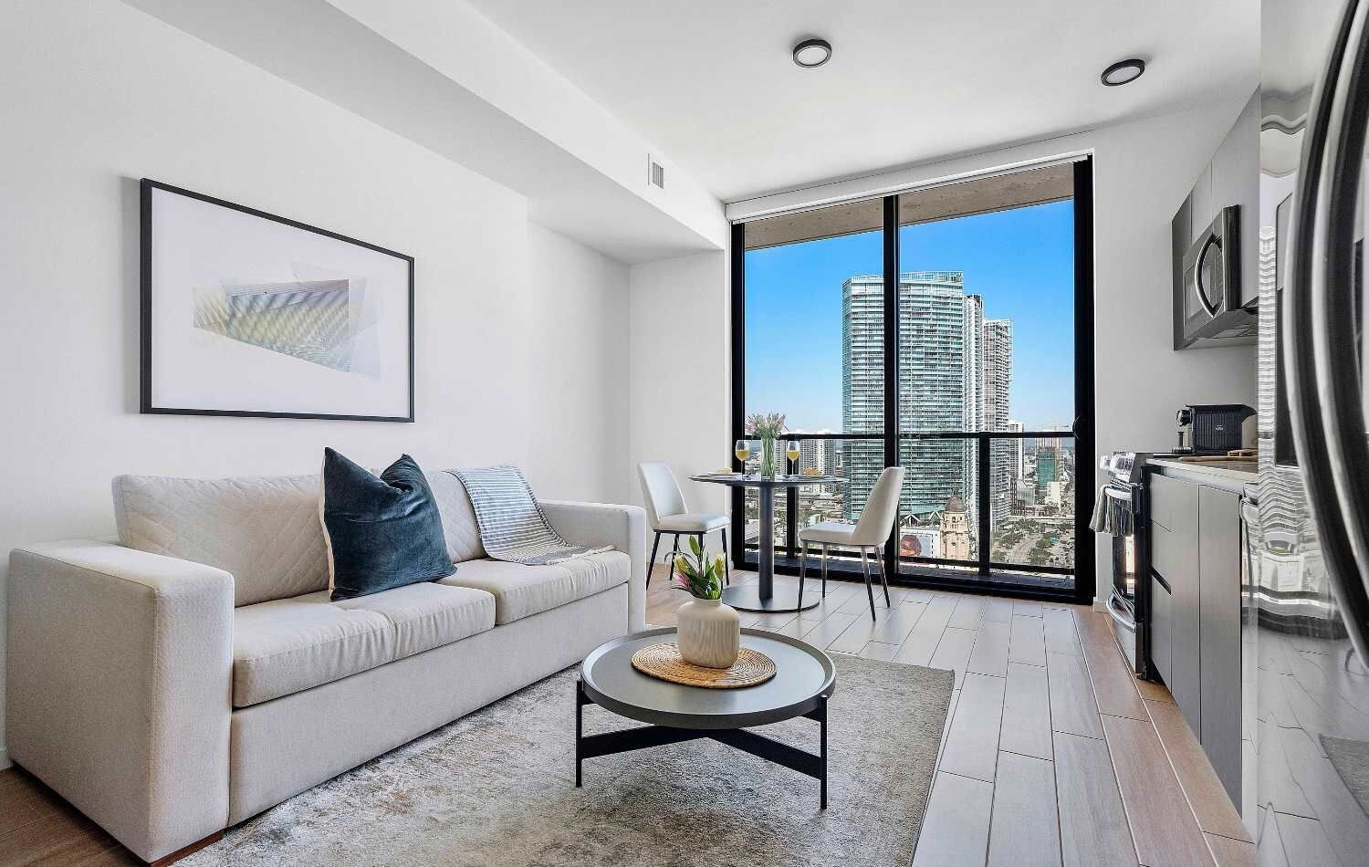 A modern living room with a sofa, coffee table, and a view of a cityscape through large windows.