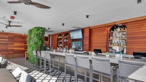A bar area with a high ceiling, ceiling fans, a TV screen, and a variety of bottles on shelves.