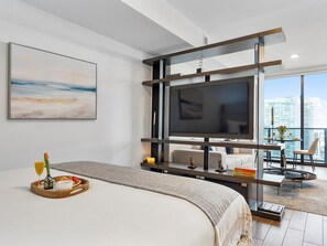 A modern hotel room with a large bed, a TV mounted on a wooden structure, a balcony with a dining table and chairs, and a city view.