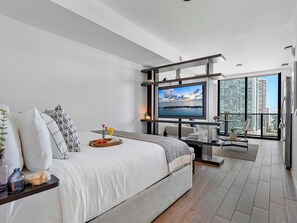 A modern hotel room with a large bed, a TV, a balcony with a city view, and a dining area.