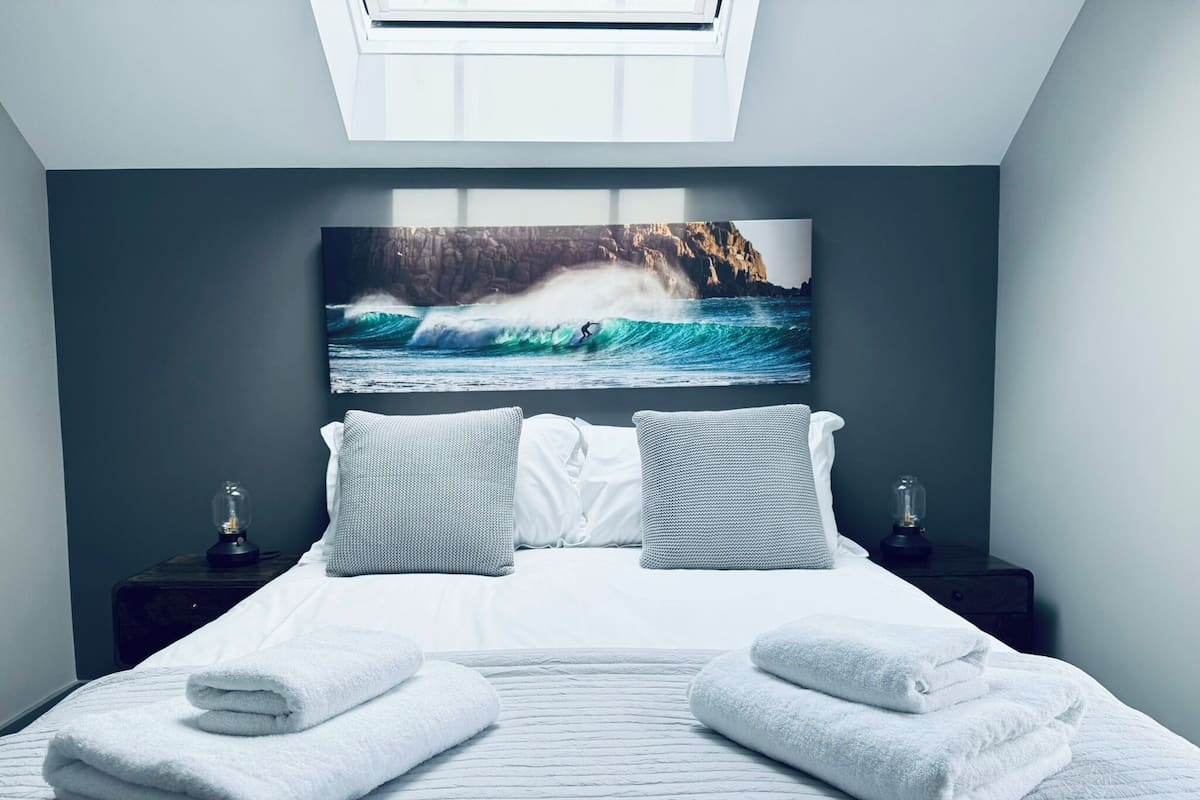 Un dormitorio con una cama grande, dos mesitas de noche, un cuadro colgado en la pared de una ola y un tragaluz.