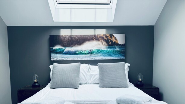 A bedroom with a large bed, two bedside tables, a wall-mounted painting of a wave, and a skylight above.