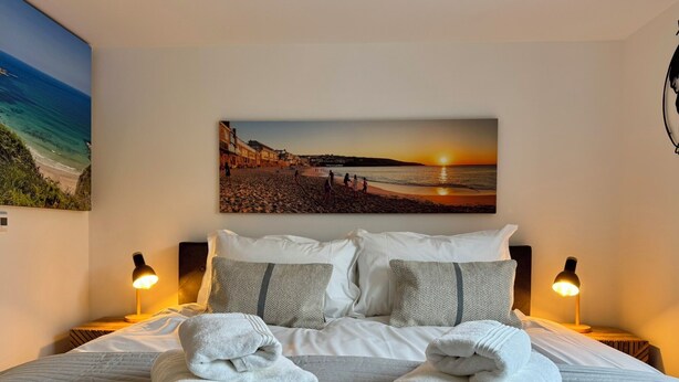 Un lit bien fait, avec du linge de lit blanc et deux serviettes pliées, une affiche représentant un coucher de soleil sur la plage au mur, et deux lampes de chevet.
