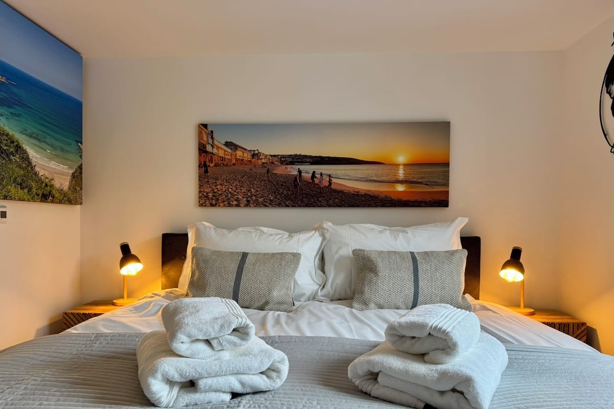 Una cama bien tendida con sábanas blancas y dos toallas dobladas, con una vista a la playa al atardecer en la pared, y dos lámparas de noche.