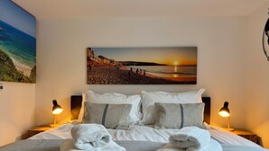 A neatly made bed with white linens and two folded towels, a sunset beach scene on the wall, and two bedside lamps.