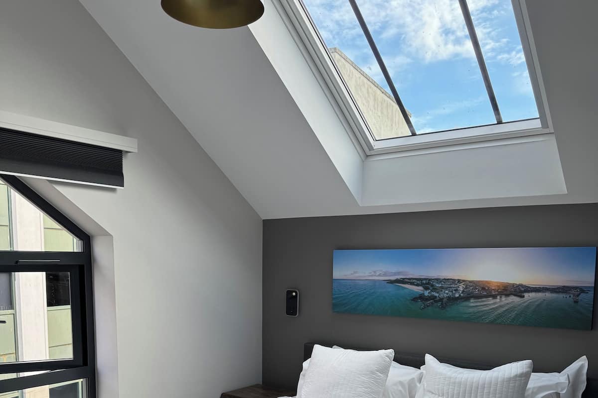 Un dormitorio moderno con una cama grande, ropa de cama blanca y vistas a un paisaje costero a través de un tragaluz.