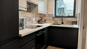 Une cuisine moderne avec des meubles de couleur sombre, un plan de travail blanc et une fenêtre donnant sur un immeuble.