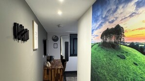 Un couloir avec un plancher en bois, un coin repas avec une table et des chaises en bois, une poubelle et une grande fresque murale représentant un paysage avec des arbres et un coucher de soleil.