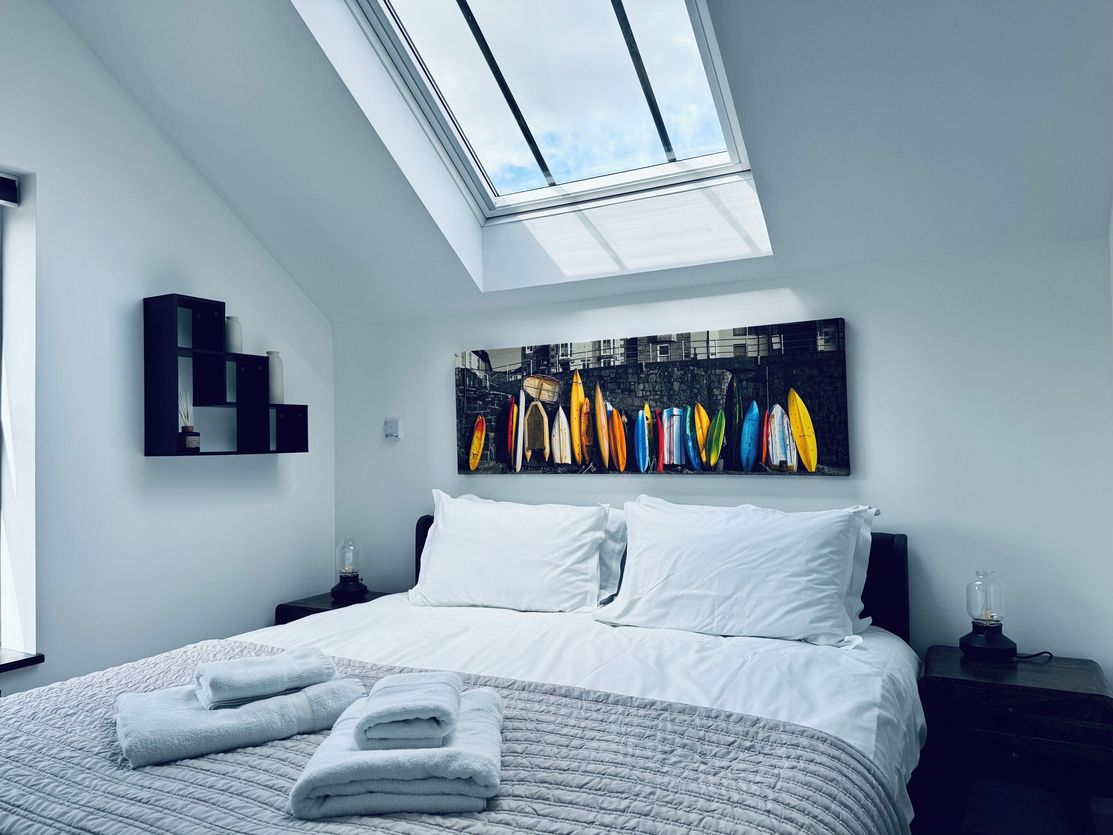 A modern bedroom with a skylight, a bed with white linens, a wall-mounted shelf, and a colorful surfboard artwork.