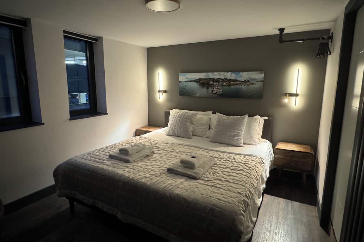 Un dormitorio ordenado con una cama grande, dos mesitas de noche, una ventana y un cuadro en la pared.