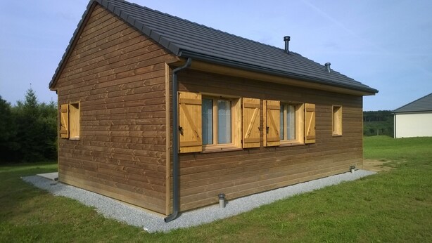 Una cabaña de madera con techo a dos aguas, ventanas amplias con contraventanas y base de grava.