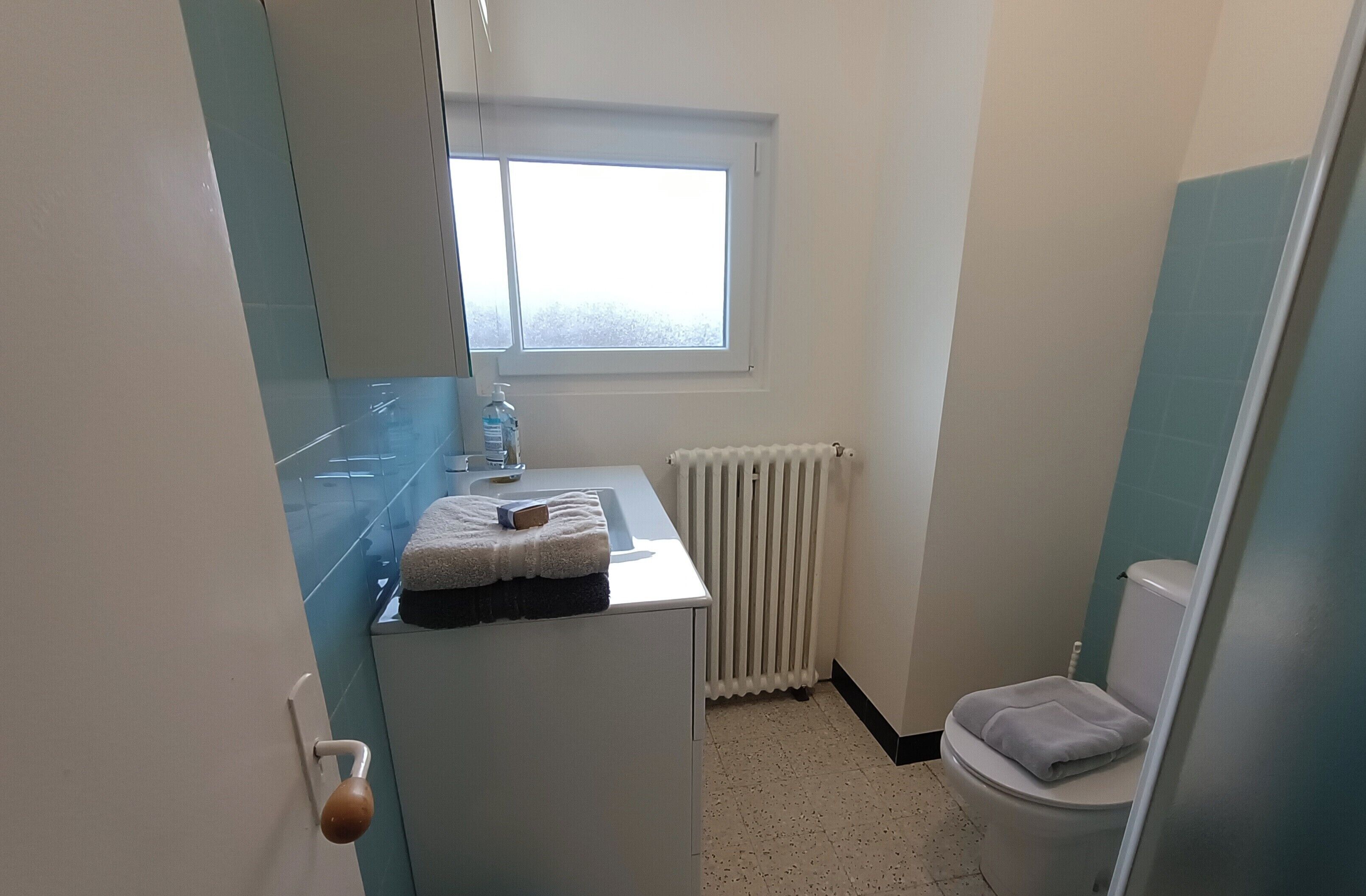 Une petite salle de bain avec une fenêtre, un radiateur, des toilettes et un lavabo sur lequel est posée une serviette.