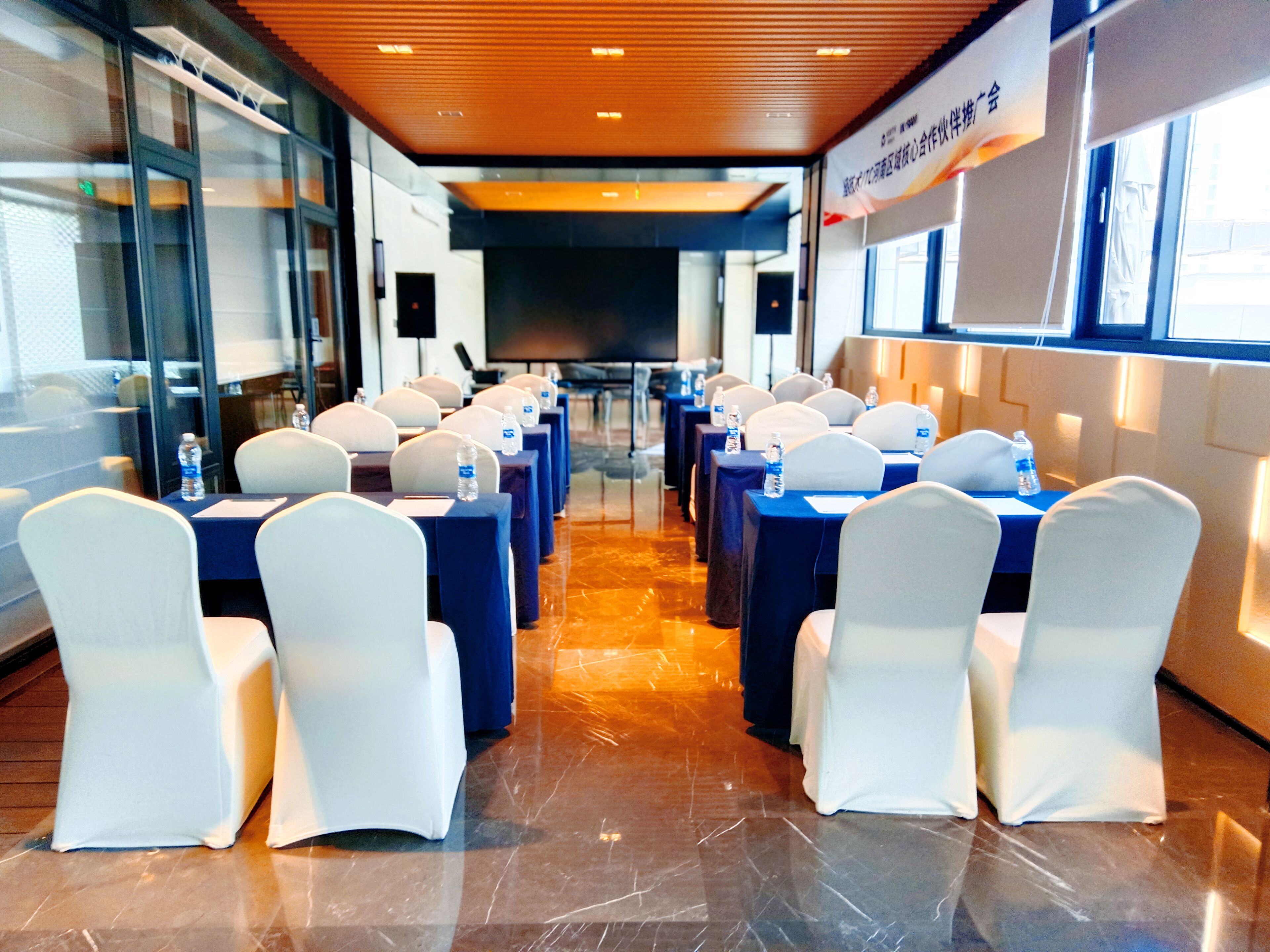 A conference room with rows of tables and chairs, a large screen, and a glass wall on one side.