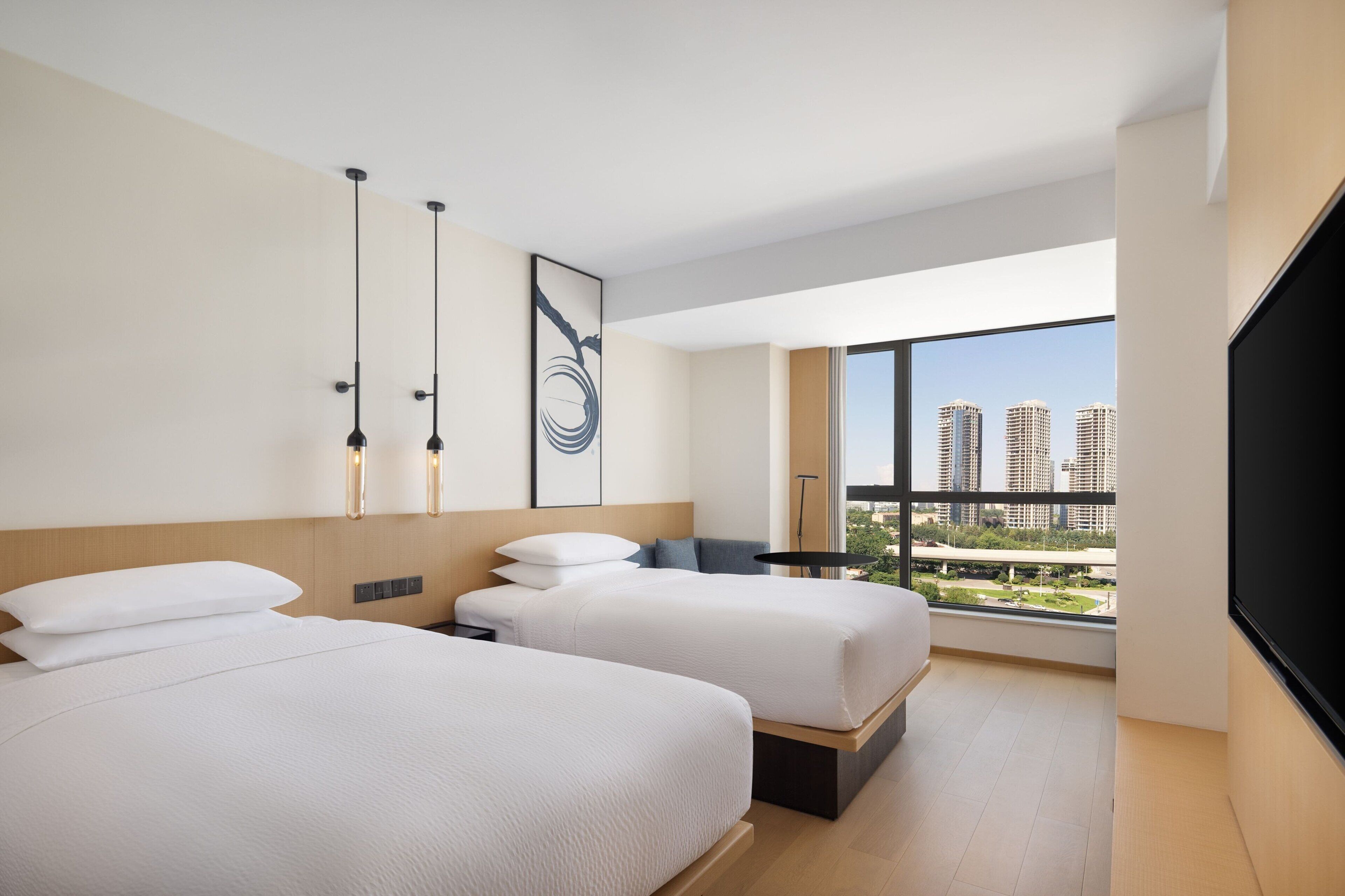 A modern hotel room with two beds, a large window with a city view, and a flat-screen TV.
