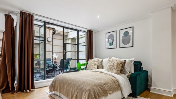 Une chambre avec une grande fenêtre, un lit recouvert d’une couverture beige, un fauteuil vert et deux tableaux encadrés accrochés au mur.