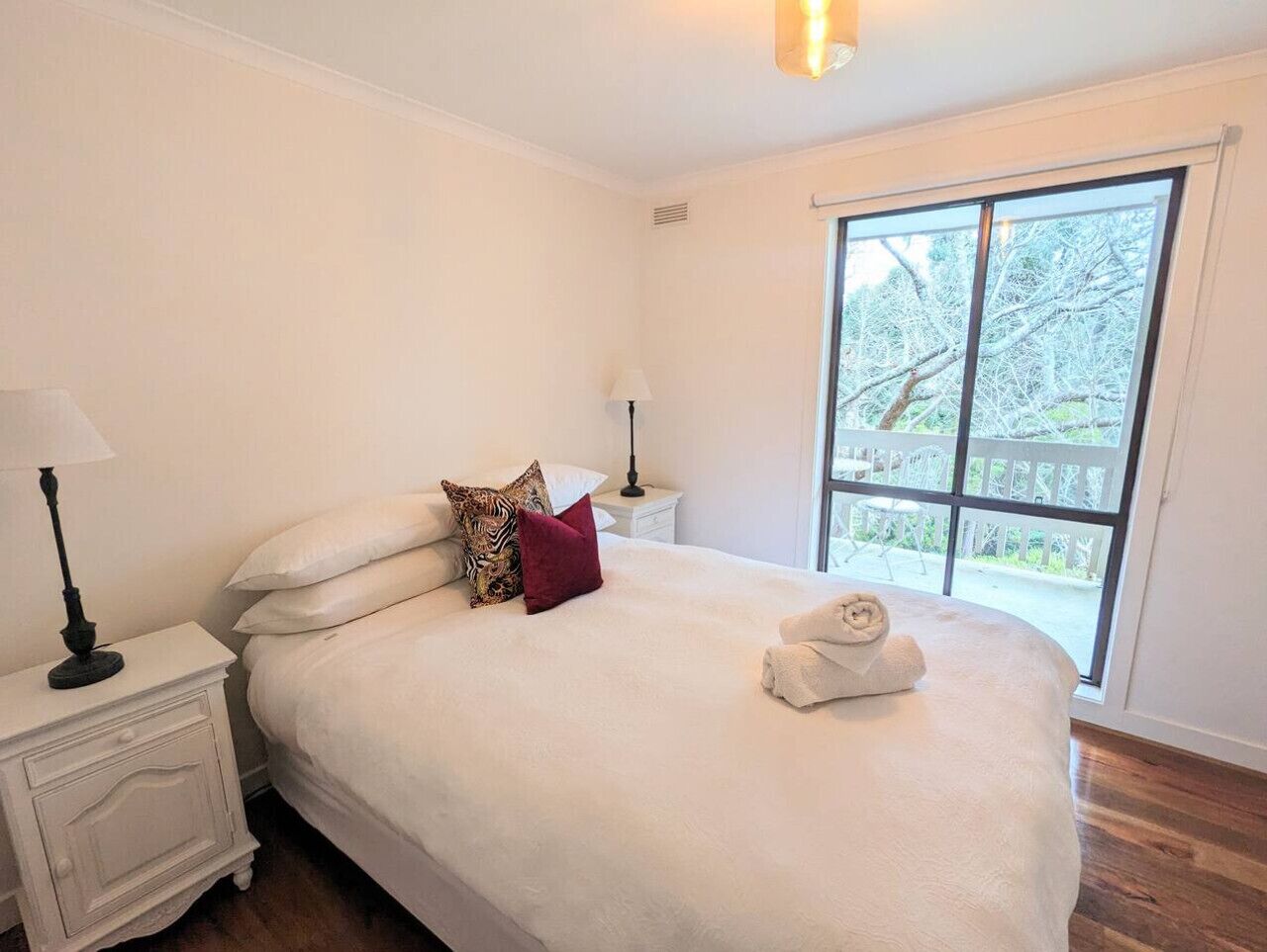 Una cama bien hecha con cubrecama blanco y una toalla enrollada, una mesita de noche con lámpara, y una ventana con vista a árboles.