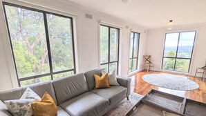 Un salon moderne avec un canapé d’angle gris, un tapis à motifs et une table basse sur laquelle est posé un plateau. De grandes fenêtres offrent une vue sur les arbres et un paysage lointain.