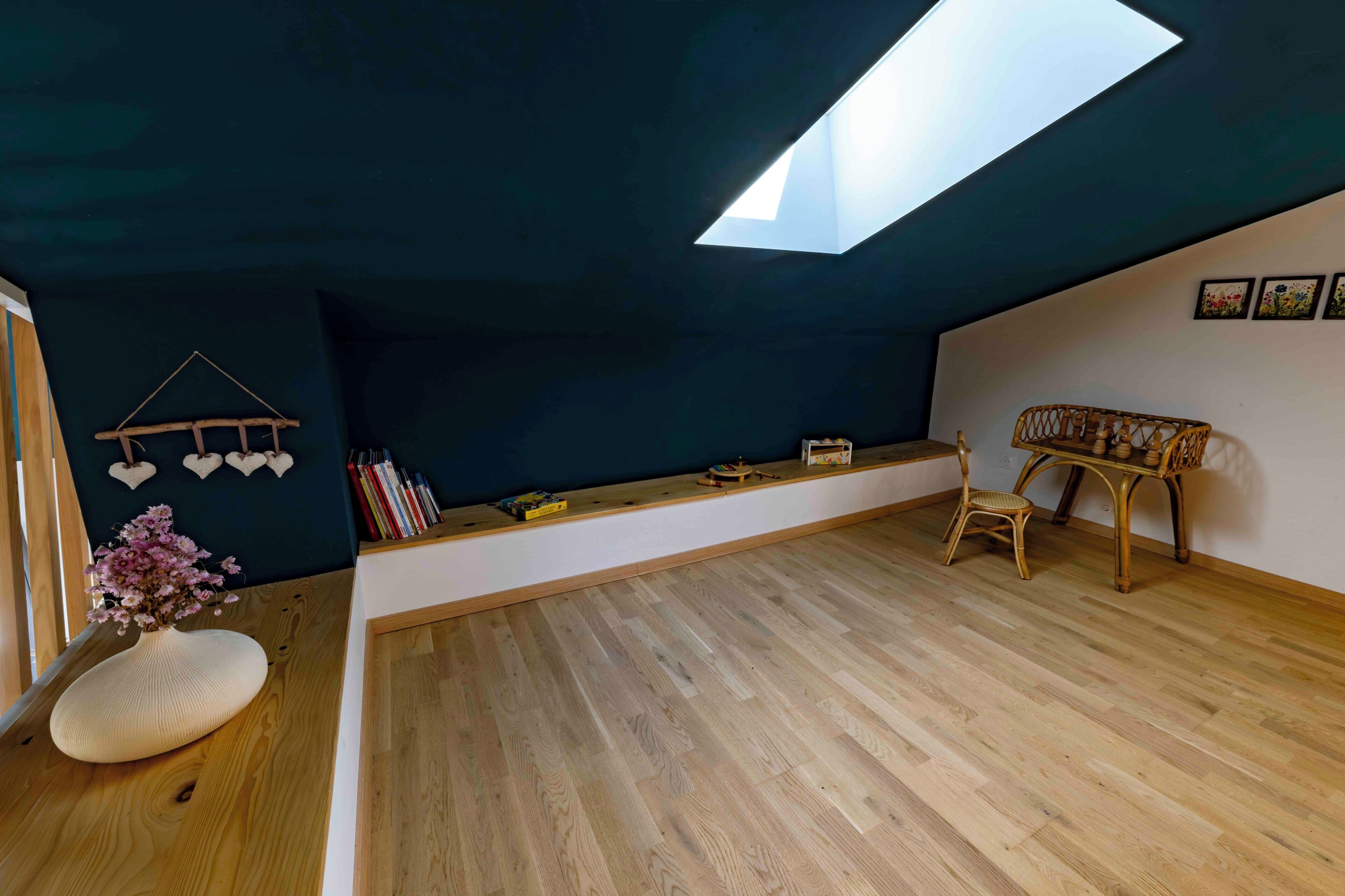 A room with a sloped ceiling, a skylight, wooden flooring, a white bench, a wicker chair, and a wall-mounted shelf with books and decorative items.