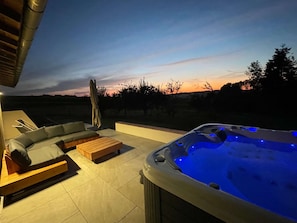 Un bain à remous avec des lumières bleues, un canapé d’angle et un banc en bois sur une terrasse offrant une vue sur le coucher de soleil.