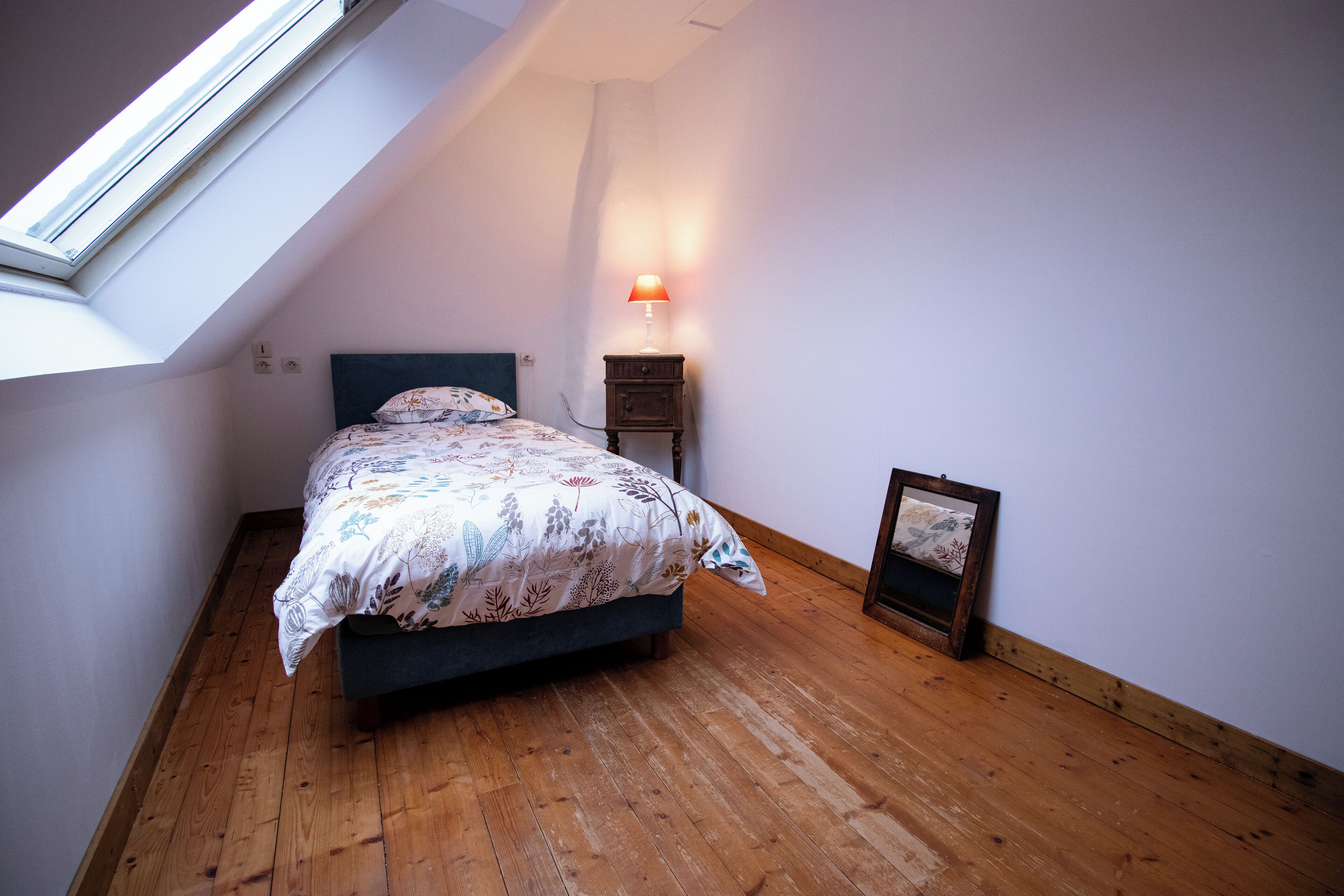 Une chambre avec un plafond en pente, un lit recouvert d’une literie à motifs floraux, une table de chevet avec une lampe et un tableau encadré accroché au mur.