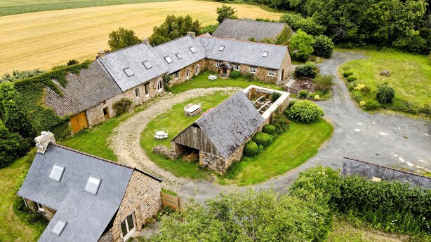 Une ferme en pierre dotée de nombreuses pièces, une cour centrale aménagée avec des sièges et un champ agricole qui l’entoure.