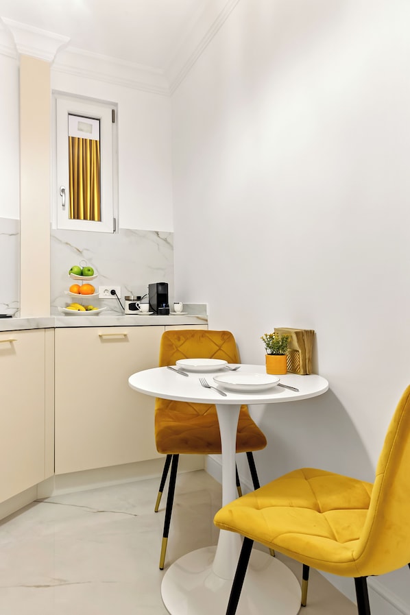A compact kitchen with a white marble countertop, a coffee machine, and a small dining area with a yellow chair and a white table.