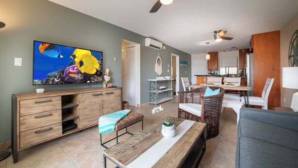 A modern living room with a flat-screen TV, wooden furniture, a dining area, and a kitchen.