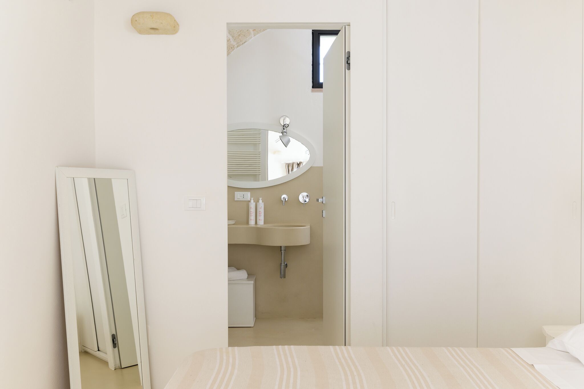 A bedroom with a bed, a white wardrobe, a bathroom with a sink and mirror, and a door leading to another room.