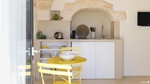 Une cuisine avec une arche en pierre, des meubles blancs, une table jaune et un saladier posé dessus.
