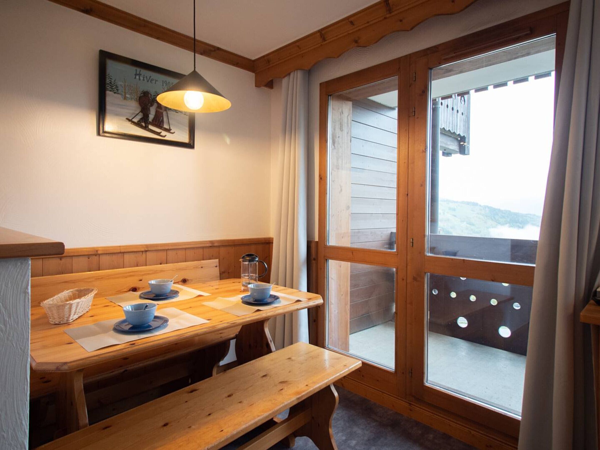 A cozy dining area with a wooden table set for two, a bench, and a large window with a view of a snowy landscape.