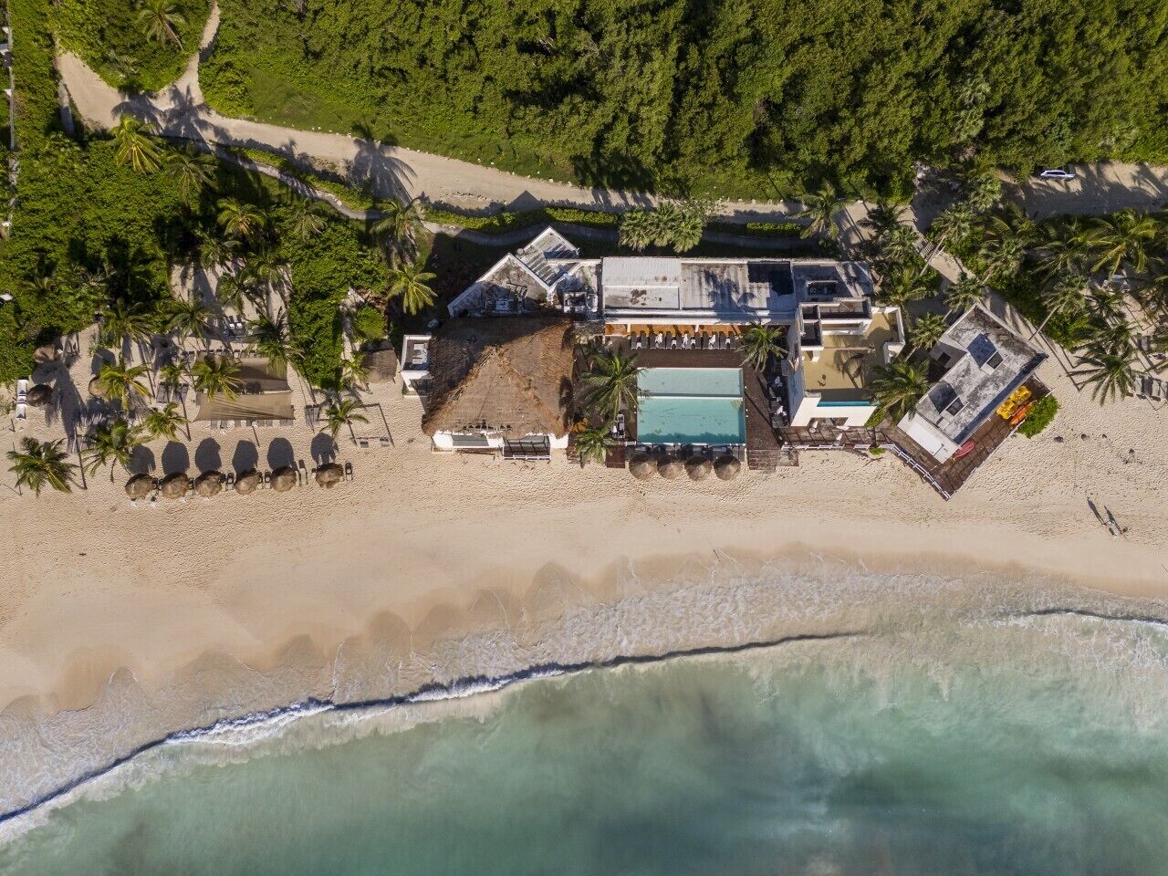 Vista aérea de un resort frente a la playa, con piscina, varias construcciones y palmeras.