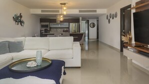 Un soggiorno moderno con un divano bianco, un tavolino da caffè con oggetti decorativi, una TV su un supporto in legno e una cucina sullo sfondo.