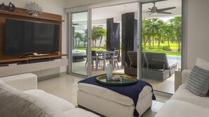 Un soggiorno moderno con una TV a schermo piatto, divani bianchi, un tavolino da caffè con un oggetto decorativo e una porta scorrevole in vetro che dà su un'area esterna con piscina e vegetazione.