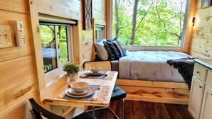 Een houten blokhutinterieur met een eettafel, stoelen, een bed met kussens, een klein raam en uitzicht op bomen.