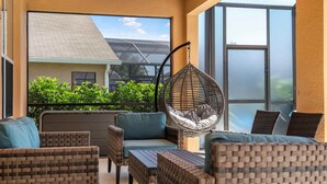 A balcony with wicker furniture, including a hanging chair and cushioned seating, overlooking a building with a sloped roof and greenery. - Avatar | 2.5 Mi Disney | Pool | Game Room 8510 (Kissimmee)