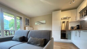 Una sala de estar moderna con un sofá gris, piso de madera y una cocina con gabinetes blancos y electrodomésticos de acero inoxidable.
